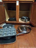 Photo showing muffin pan, bundt pan, square skillet, and large non-stick skillet with lid on wooden floor in front of open cabinet.
