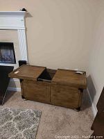 Wooden storage chest with dual hinged lids open showing interior space. Brown wood with visible grain, set against beige wall and carpet.