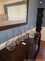 Seven glass bar ware items including pitchers, decanters, and bottle stoppers displayed on a dark wood console table against a blue wall beneath a large rectangular mirror.