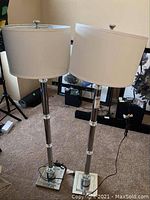 Two floor lamps standing side by side on carpet near wall with artwork behind. Lamps have metal poles with acrylic accents and white cylindrical shades. Both cords visible on floor.