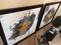 Two large framed abstract art pieces with blue and gold splash designs leaning against a wall on a carpeted floor.