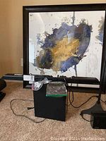 Samsung sound bar, subwoofer, and remote control shown together with framed artwork behind on carpeted floor.