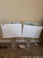 Photo of two glass block table lamps with white rectangular fabric shades and light bulbs included, shown on carpeted floor against wall.
