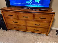 Front view of wooden dresser with 7 drawers showing medium-tone wood and black metal handles, placed on carpeted floor.