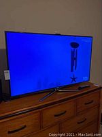 Front view of TV powered on, showing screen, V-shaped stand on dresser