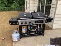 Photo of the black Pit Boss grill smoker with propane tank positioned outside on a concrete slab near a house. Shows full unit and attached propane tank.