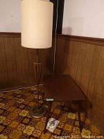 Photo showing the wooden rectangular table and the tall floor lamp together in a corner with wood panel walls and patterned floor tiles.
