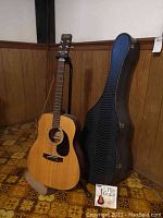 Full view of acoustic guitar on stand beside hard shell case and instructional CD