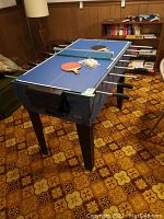 Full view of Cooper multi-game table showing foosball rods, ping pong paddles and balls on top, in wood-paneled room.