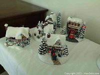 Five-piece set of snow-covered Christmas village houses with trees and figures, including First Responders house with red fire truck, Childhood Home cottage with decorated tree and snowman, displaying each piece lit with battery power.
