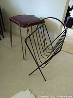 Mid century stool with maroon vinyl padded seat and hairpin legs next to black wire magazine rack with curved top and angled legs.