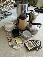 Overall lot contents showing coffee percolators, kettle, thermal carafe, mugs, placemats, and coasters arranged on carpet.
