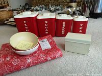 Full lot view showing four red plastic cannisters labeled Flour, Sugar, Coffee, Tea, white metal recipe box, two white bowls, and red printed plastic tablecloth