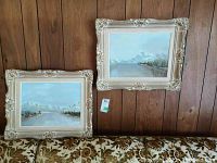 Two framed acrylic paintings on board depicting mountain lake landscapes, hung on wood-paneled wall above couch.