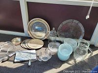Photo of the entire lot showing glass bowls stacked, large glass tray, glass pitcher, covered dish, marble cheese tray with spreader, silverplate tray, trivet, and tongs