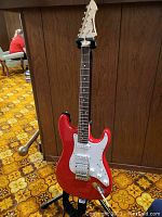 Full view of red and white electric guitar on black Hercules stand against wood panel background.