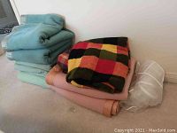 Folded pastel blue, pastel green, pink blankets in synthetic material with a multi-colored checkered wool blanket on top, and two white zippered storage bags next to the pink blanket.