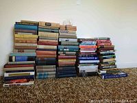 Stack of about 70 hardcover books arranged in multiple piles showing variety of sizes and colored cloth and paper bindings on carpeted floor