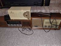General view of three mid-century radios placed on carpet in front of a fireplace, showing the three radios including a tan plastic General Electric, a wooden Onkyo, and a wooden Panasonic radio with cords visible.