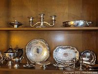 Two shelves filled with various silverplate items including trays, teapot, serving pieces, candelabra, and candle holders arranged neatly.