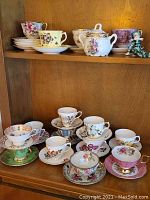 Shelf with fifteen assorted porcelain cups and saucers arranged in three rows along with a broken leg porcelain figurine in green and white clothing.
