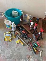 Wide view of lot items including teal bucket, windshield washer fluid containers, hydraulic jack, jack stands, tools and hardware pieces on concrete floor.