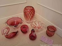 Collection of cranberry glass items including swan, fruit figurines, two bowls, and a vase shown together on carpeted floor against wall corner.