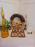 Tall yellow glass decanters, grapevine wreath with artificial sunflowers, four framed dried flower art, brass hurricane candle holder, and white wax bowl with pressed botanicals on doily.