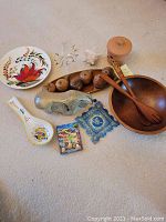 Full view of all items included in the lot arranged on a neutral floor: wood salad bowl with wooden utensils, clay garlic pot, ceramic bull, glass bottle with gold accents, ceramic and decorated dishes, sea shell, and small decorative clay pears.