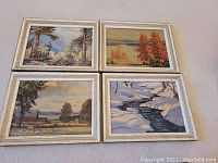 Four framed oilettes arranged on a flat surface, showing different nature scenes including forest, autumn trees, lake with farmhouse, and winter creek
