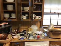 Wide view of the kitchen counter showing various stacked stoneware plates, mugs in cabinet and on counter, assorted glassware, and the edge of microwave and vintage metal pitcher