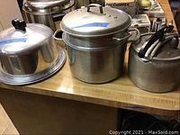 Large metal cooking pots with lids, cake keeper, kettle, and pressure cooker on wooden surface