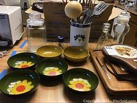 Photo showing six vintage plastic bowls with floral designs, two glass jars with tops, ceramic utensil holder with utensils, and wooden cheese boards on table