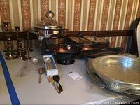 Overall view of silver plate platters, trays, bowl, serving pieces and 8 brass candle holders arranged on a table.