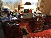 Photo of large wooden desk with multiple drawers covered with assorted office supplies such as file folders, calculators, pens, tape dispensers, an adding machine, and various papers.