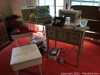 Wide view of sewing machine on table, chair with torn seat, sewing basket, and box on top, with parts and accessories around.
