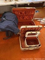 Three opened matching cream-colored vintage Travel Joy suitcases with orange ruched inner lining, compartments, and clasps. Gray garment bag nearby.