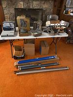 Photo shows a white plastic tabletop with several vintage film projectors including Bell and Howell and others, a set of metal lighting fixtures with multiple bulbs, multiple cardboard boxes possibly containing accessories or components, and a group of metal roll-up projector screens laid out on the floor in front of the table.