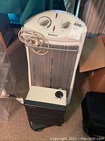 Holmes tower heater with visible front grill, dual knobs and power cord coiled on side. Second black Tatung heater positioned in front on floor.