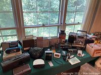 Table displaying an assortment of radios, clocks, microphone, recorders, cables, and CD players arranged in front of large window.