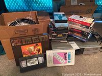 Boxed electronics and various VHS and DVD media items placed on carpeted floor next to cardboard box.