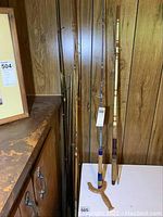 Photo showing assortment of fishing rods leaning against a wood-paneled wall, some with cork handles and others with synthetic grips. No reels present.