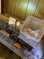 View of American Flyer black steam locomotive, tender, red caboose, metal curved train tracks, and plastic bin with wooden accessories on a table.