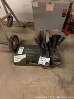 Photo shows pair of worn Pankow size 11 boots standing on the floor beside a metal German hand grenade case that is military green with German text and stamps.