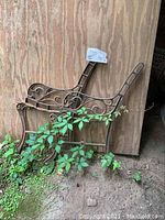 Pair of cast-iron bench ends leaning against wood wall with some plants around the base.