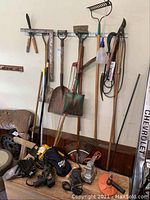 Wide view of hanging and standing garden tools including shovels, rake, pruner, brooms, power tools on floor including drills and sander, chainsaw and other hand tools; some handles show damage.