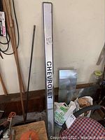 Full view of the long silver metal tailgate panel with 'CHEVROLET' embossed in black letters, leaning against wall above garage items.