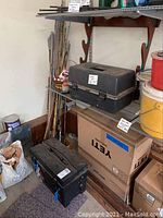 Fishing rods stored vertically in a corner next to a shelf with two plastic tackle boxes on top and Yeti cardboard boxes underneath. Yellow and red bait buckets are nearby.