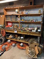 Full view of the workbench with shelves holding multiple small parts organizers, paint containers, parts boxes, hand tools, and various hardware. Coiled extension cords and other wires on floor and lower shelf.