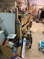 Wide view of basement workshop showing sawhorses, wooden planks, boxes with tools and extension cords, and other hardware parts scattered on shelves.
